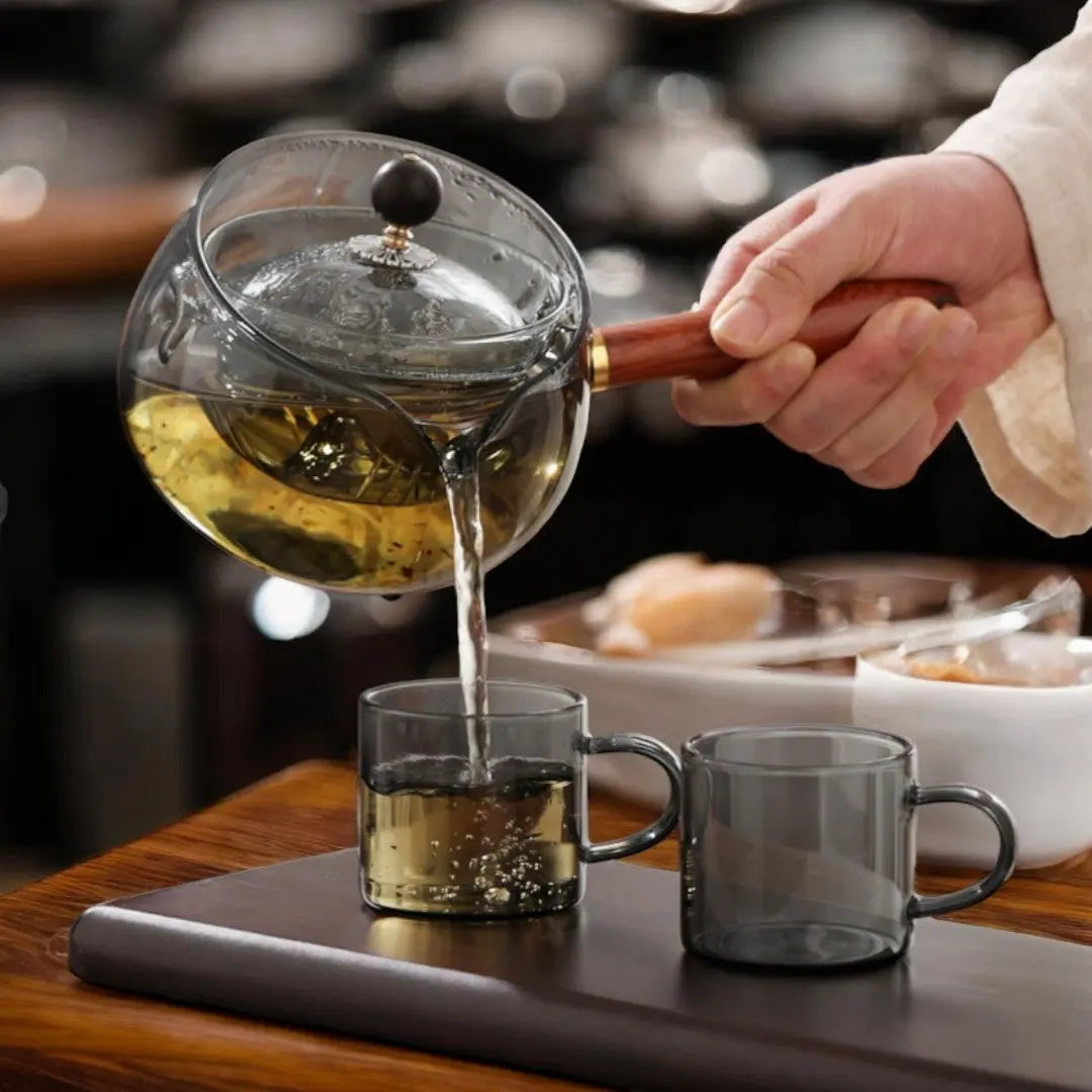 Pouring amber tea from smoked gray glass teapot into matching cup set