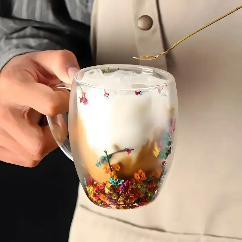 Hand holding insulated glass mug with layered drink and colorful suspended dried flowers