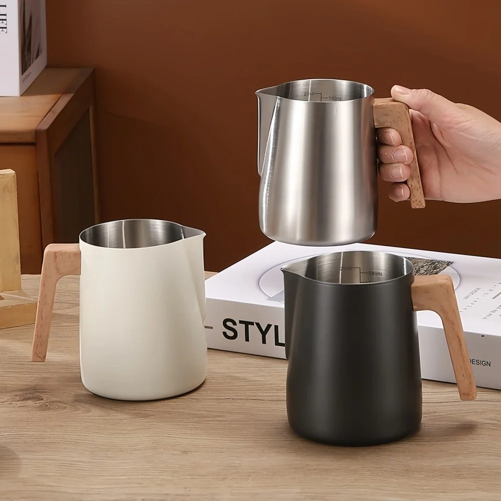 Three matte cream, black and stainless steel barista milk pitchers with wooden handles on wood surface