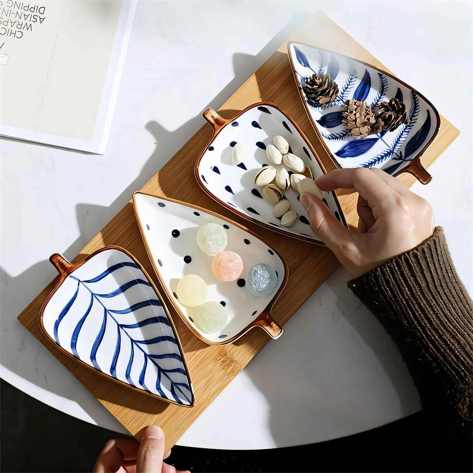 Top view of four blue patterned ceramic leaf dishes on bamboo tray with pistachios