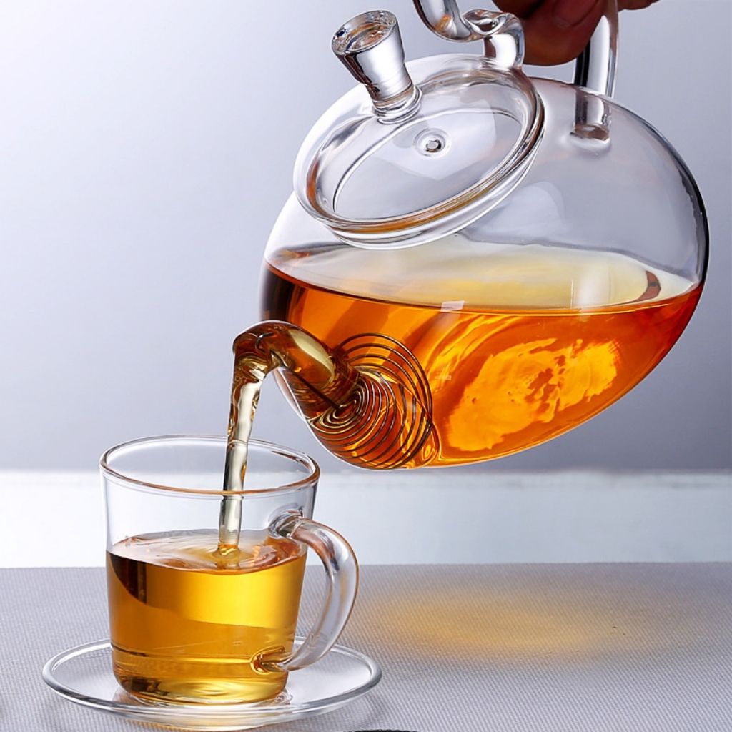 Pouring amber tea from clear glass teapot with internal coil filter into cup