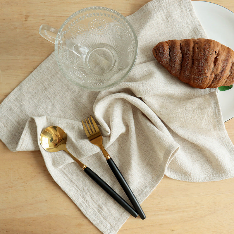 Beige cotton linen dining napkin styled with croissant and gold cutlery on wood