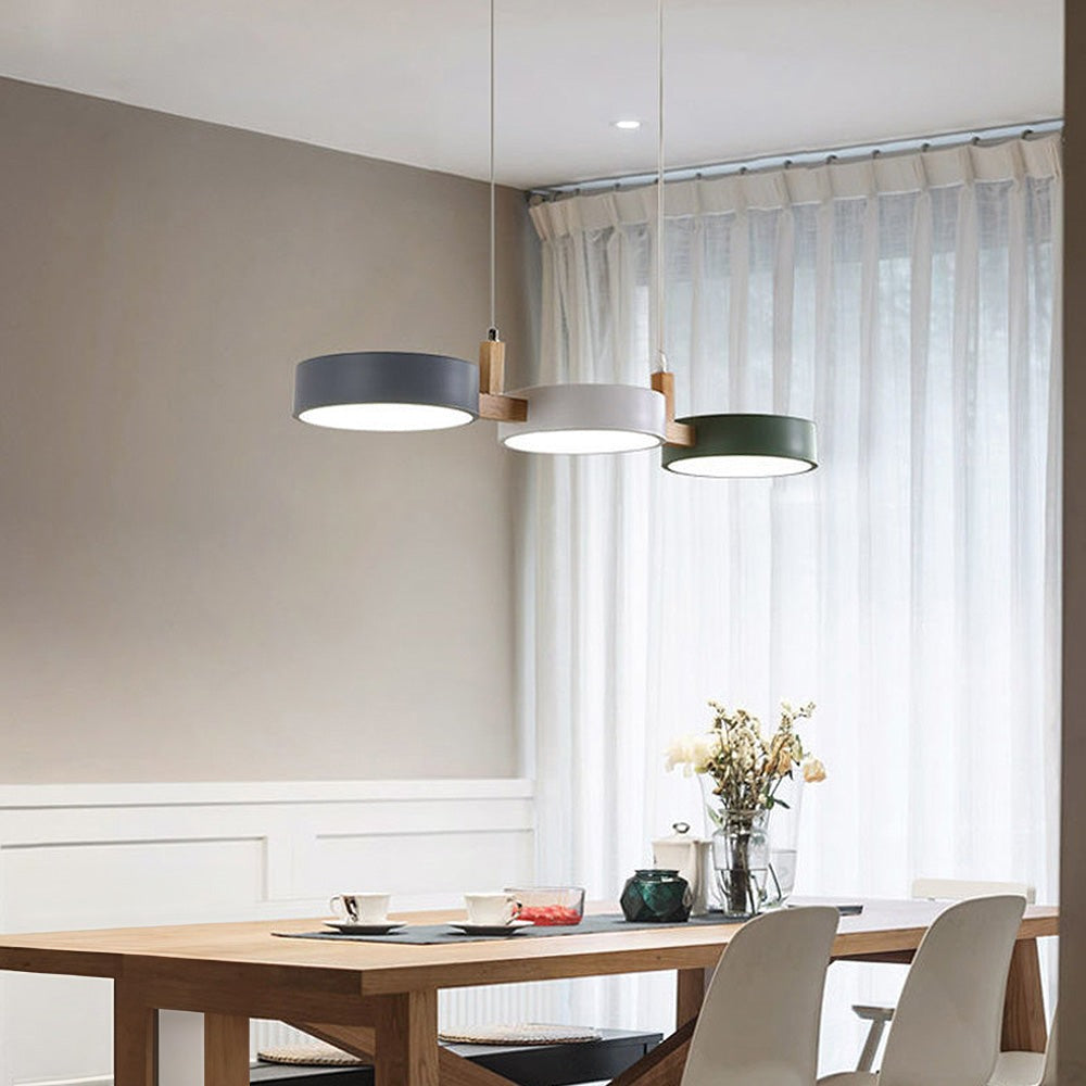 Maintained House Nordic Wood Drum LED Pendant - Gray, White and Green drums over a wooden dining table by a window